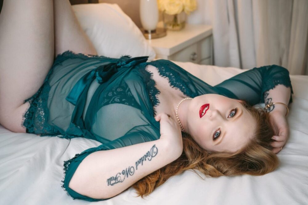 beautiful red headed woman laying on her back on the boudoir studio bed wearing a dark green sheer robe