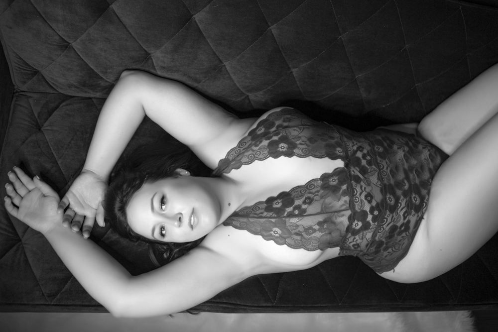 black and white photo of curvy woman in lacy bodysuit laying on her back with her arms over her head on the boudoir studio couch