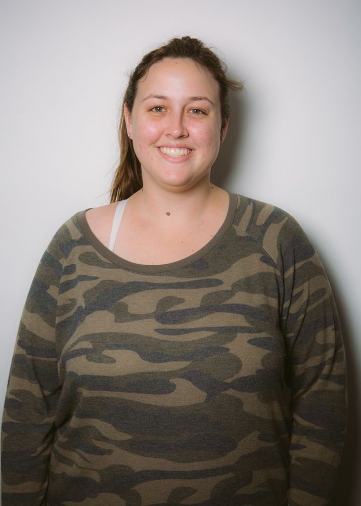 A woman in a camouflage shirt smiling in front of a white wall.