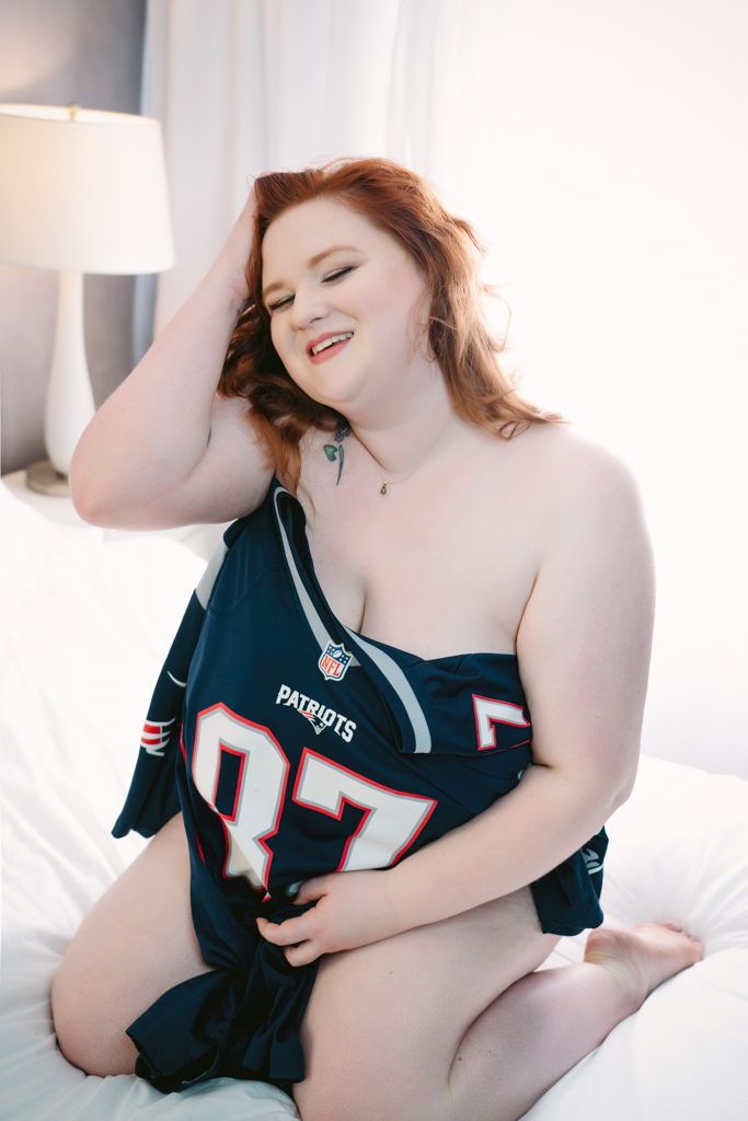 A woman wearing a new england patriots jersey on a bed.