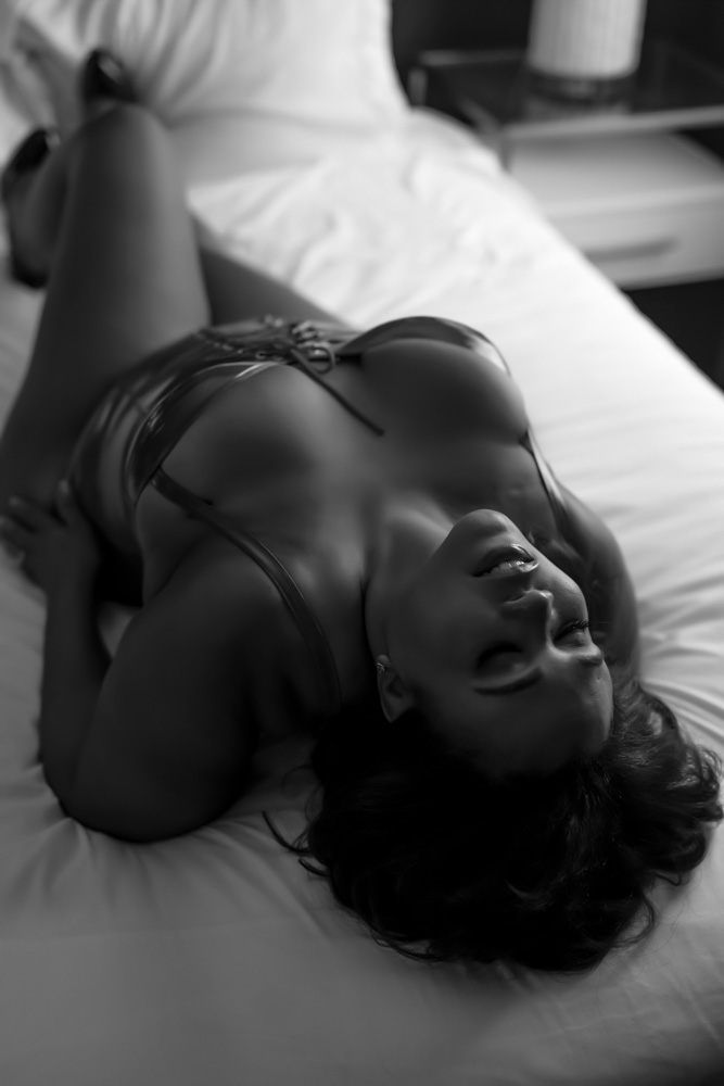 black and white photo of a curvy woman laying on her back with her head tilted back and eyes closed with her hands on her hips on the boudoir studio bed